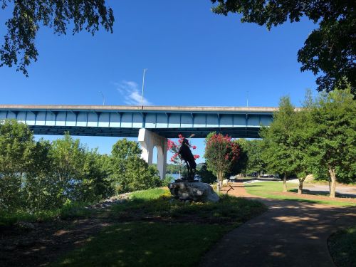 A statue of a horse and rider stands on a rock beneath a blue bridge, surrounded by trees and greenery.