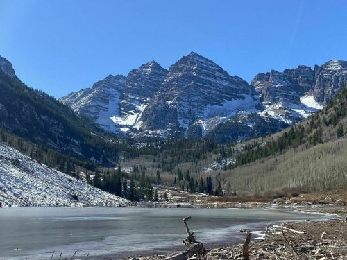 Snow-capped mountains rise above a frozen lake, surrounded by evergreen trees and a clear blue sky.