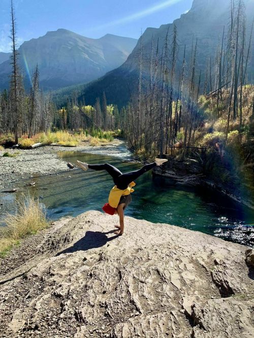 A person performs a handstand on a rock by a river, surrounded by mountains and autumn foliage.