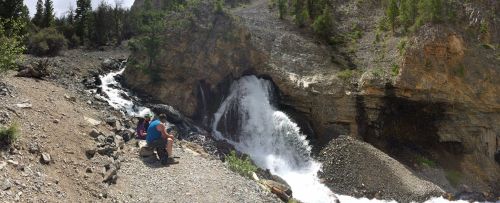 A panoramic view of a waterfall cascading down rocky terrain, with two people sitting nearby, enjoying the scenery.