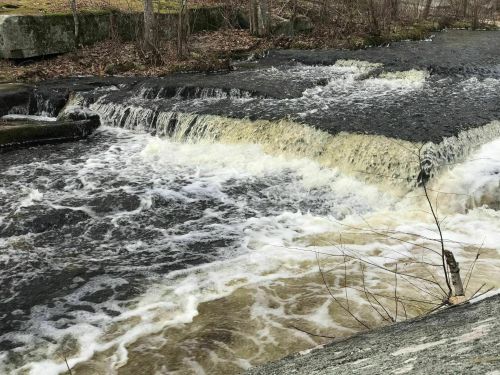 A flowing river cascades over rocks, creating white frothy water against a backdrop of bare trees and earthy tones.