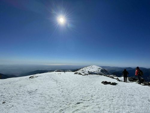 Two hikers on a snowy mountain peak under a bright sun, with a clear blue sky and distant hills in the background.
