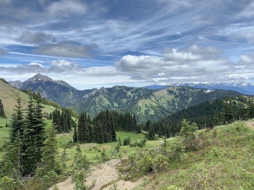 A panoramic view of lush green mountains under a partly cloudy sky, with dense forests and distant peaks.