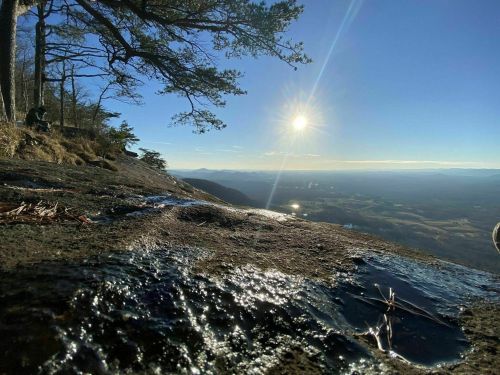 A sunlit landscape from a rocky viewpoint, with trees and distant mountains under a clear blue sky.