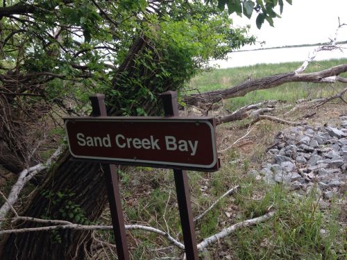 A sign reading "Sand Creek Bay" near a grassy area and water, surrounded by trees and rocks.