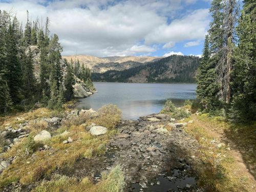 A serene lake surrounded by rocky shores and lush trees, with mountains in the background under a partly cloudy sky.