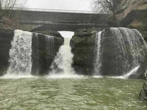 A waterfall cascades over rocky cliffs beneath a bridge, surrounded by trees and misty weather.