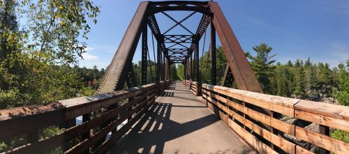 A panoramic view of a metal bridge with wooden railings, surrounded by trees and a clear blue sky.