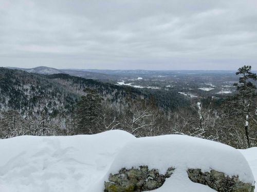 A snowy landscape view from a mountain, showcasing rolling hills and a valley under a cloudy sky.