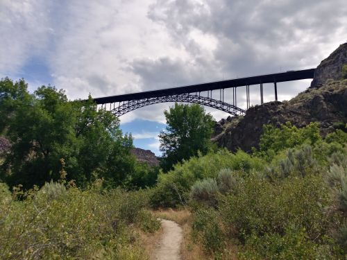 A tall bridge spans above a lush, green landscape with a dirt path leading through the vegetation under a cloudy sky.