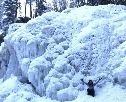 A person sits in front of a large, icy waterfall surrounded by snow-covered trees.