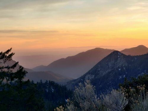Sunset over rolling mountains, with layers of peaks and valleys bathed in warm orange and purple hues.