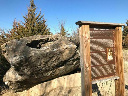 A large rock with a hole, next to an informational sign about Indiana Rock, surrounded by trees and clear blue sky.