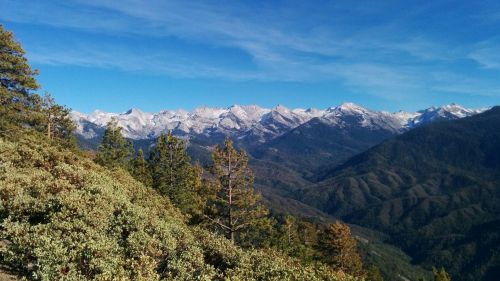 A panoramic view of snow-capped mountains under a clear blue sky, surrounded by lush green hills and trees.