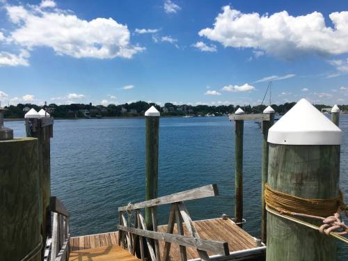 A serene waterfront view with wooden docks, posts, and a clear blue sky dotted with clouds.
