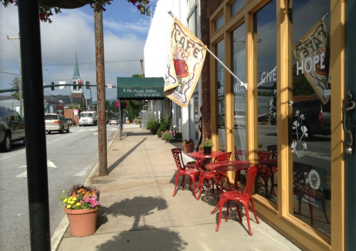 A quaint café with outdoor seating and a colorful flag, located on a sunny street with flower pots and a church spire in the background.