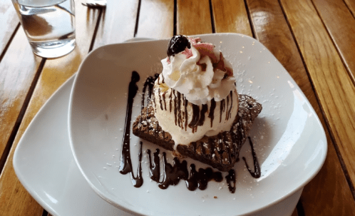 A decadent brownie topped with ice cream, whipped cream, and chocolate drizzle, served on a white plate.