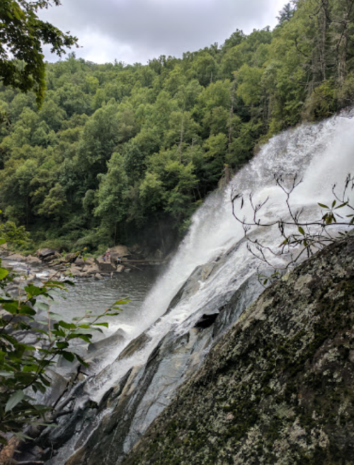 A waterfall cascades over rocky terrain, surrounded by lush green trees and a calm river below.