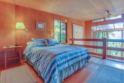 Cozy bedroom with a striped bedspread, wooden walls, large windows, and a view of trees outside.