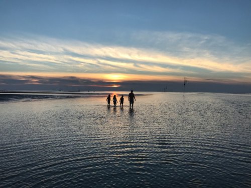 A silhouette of a person and two children walking through shallow water at sunset, with a colorful sky in the background.
