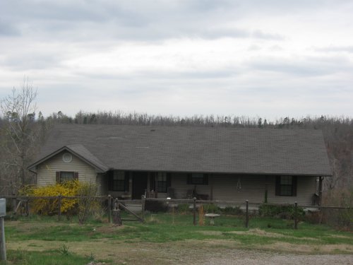 A single-story house with a gray roof, surrounded by a fence and sparse trees under a cloudy sky.