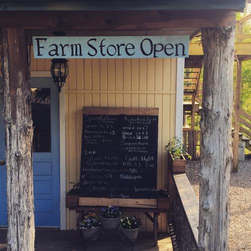 A rustic farm store entrance with a sign reading "Farm Store Open" and a chalkboard menu of available products.