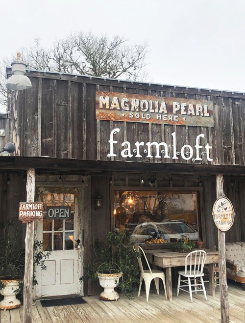 A rustic storefront with a wooden sign reading "Magnolia Pearl - Sold Here" and "farmloft," featuring an open door.