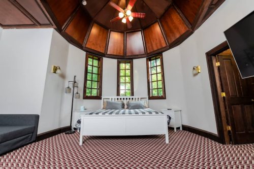 Cozy bedroom with a round wooden ceiling, large windows, a white bed, and a patterned carpet.