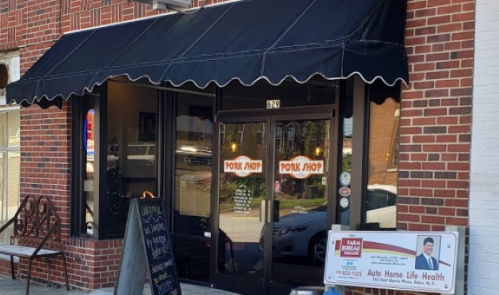 Exterior of a brick building featuring a black awning with "Pork Shop" signage and a sidewalk display board.