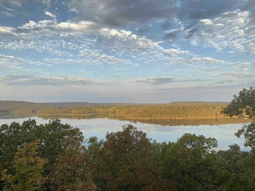 A serene landscape featuring a river surrounded by trees, with a clear sky and soft clouds reflecting on the water.