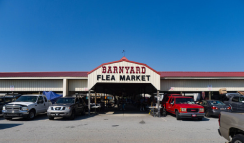 Entrance to Barnyard Flea Market with parked cars and a clear blue sky above.
