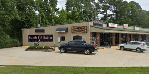A storefront with a drive-thru, featuring "Bundt Babies" signage and parked vehicles in a tree-lined area.