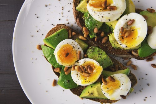 Two slices of whole grain toast topped with avocado, sliced boiled eggs, pine nuts, and black pepper on a white plate.