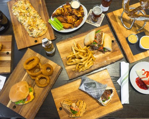 A variety of dishes on wooden boards, including sandwiches, wings, fries, onion rings, and a pretzel, with sauces.