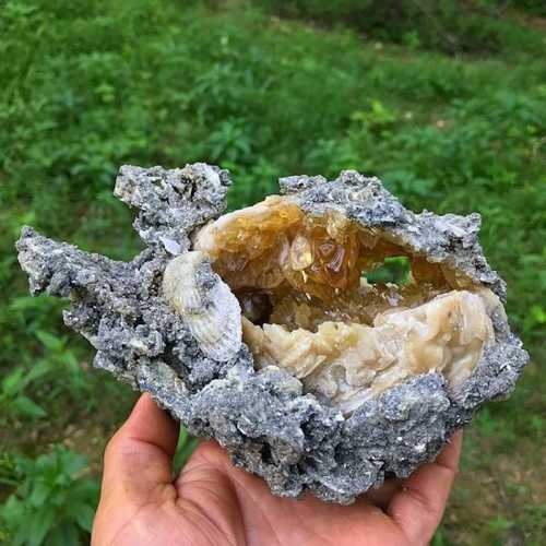 A hand holding a large, geode-like rock with a shiny, amber-colored interior and rough, gray exterior.