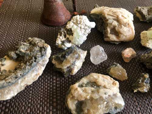 A collection of various minerals and crystals on a textured surface, with a small bell in the background.