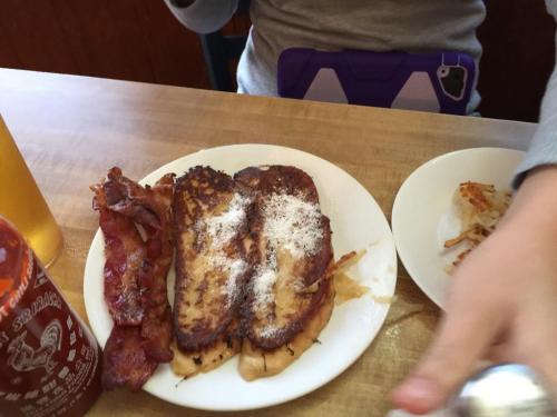 A plate with French toast dusted with powdered sugar, crispy bacon, and a side of hash browns, with a drink nearby.