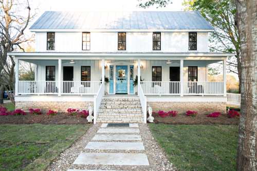 A charming white two-story house with a blue door, surrounded by greenery and pink flowers, featuring a welcoming porch.