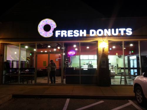A brightly lit storefront of "Fresh Donuts" at night, featuring a neon "OPEN" sign and a person standing outside.