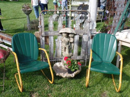 Two green chairs sit on grass in front of a decorative garden statue and a rustic fence. Flowers are in the statue's base.