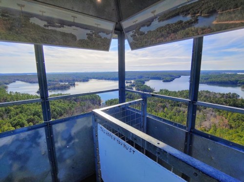 View from a lookout tower overlooking a lake surrounded by trees and rolling hills under a clear blue sky.