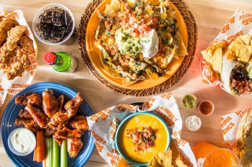 A spread of delicious food including nachos, wings, cheese dip, and drinks on a wooden table.