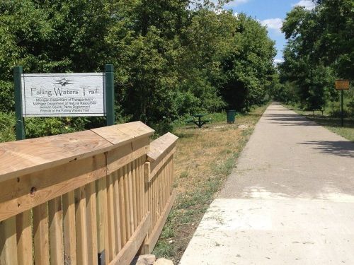 Sign for Falling Waters Trail with a paved path surrounded by greenery on a sunny day.