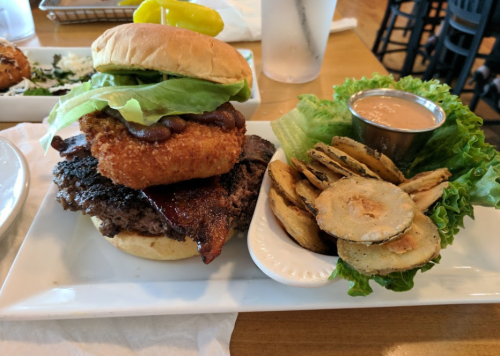 A hearty burger topped with lettuce, beans, and a crispy onion ring, served with fried pickles and dipping sauce.
