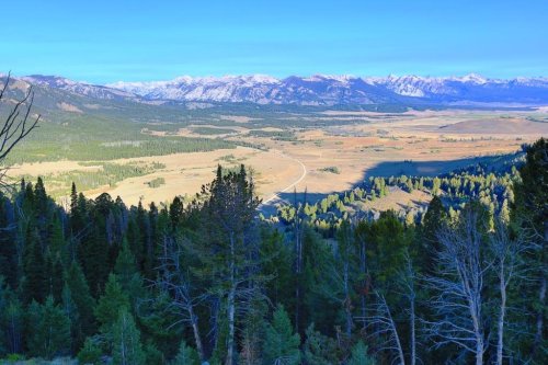 A panoramic view of a valley surrounded by mountains, with lush forests and a winding road in the foreground.