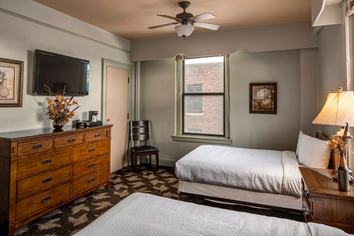Cozy hotel room with two beds, a dresser, TV, and a window, featuring warm decor and a ceiling fan.