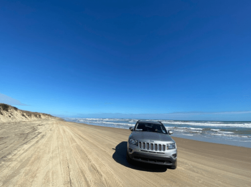 A silver SUV parked on a sandy beach with waves and a clear blue sky in the background.