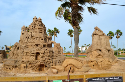 A large, intricate sandcastle with towers and details, surrounded by palm trees and a clear blue sky.