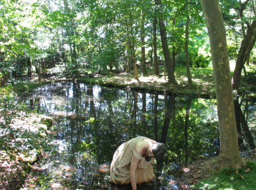 A person in a light dress bends over a serene pond surrounded by lush greenery and trees.