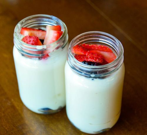 Two glass jars filled with creamy yogurt topped with sliced strawberries and blueberries, placed on a wooden surface.